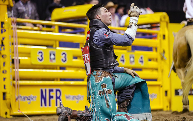 Jess Pope kneeling on one knee pointing at the sky with one hand in a rodeo arena.